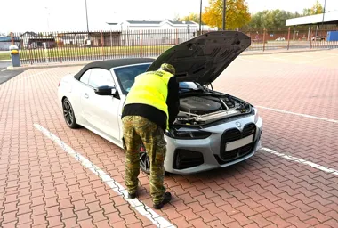 Funkcjonariusz w kamizelce odblaskowej i mundurze moro sprawdza silnik białego kabrioletu BMW na parkingu z kostką brukową.