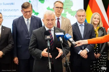 Mężczyzna w garniturze przemawia do mikrofonów mediów podczas konferencji prasowej, za nim stoją inne osoby oraz mapa i flagi.