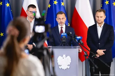 Trzech mężczyzn w garniturach podczas konferencji prasowej przy pulpicie z orłem, w tle flagi Polski i Unii Europejskiej, przed nimi kamera i mikrofony.